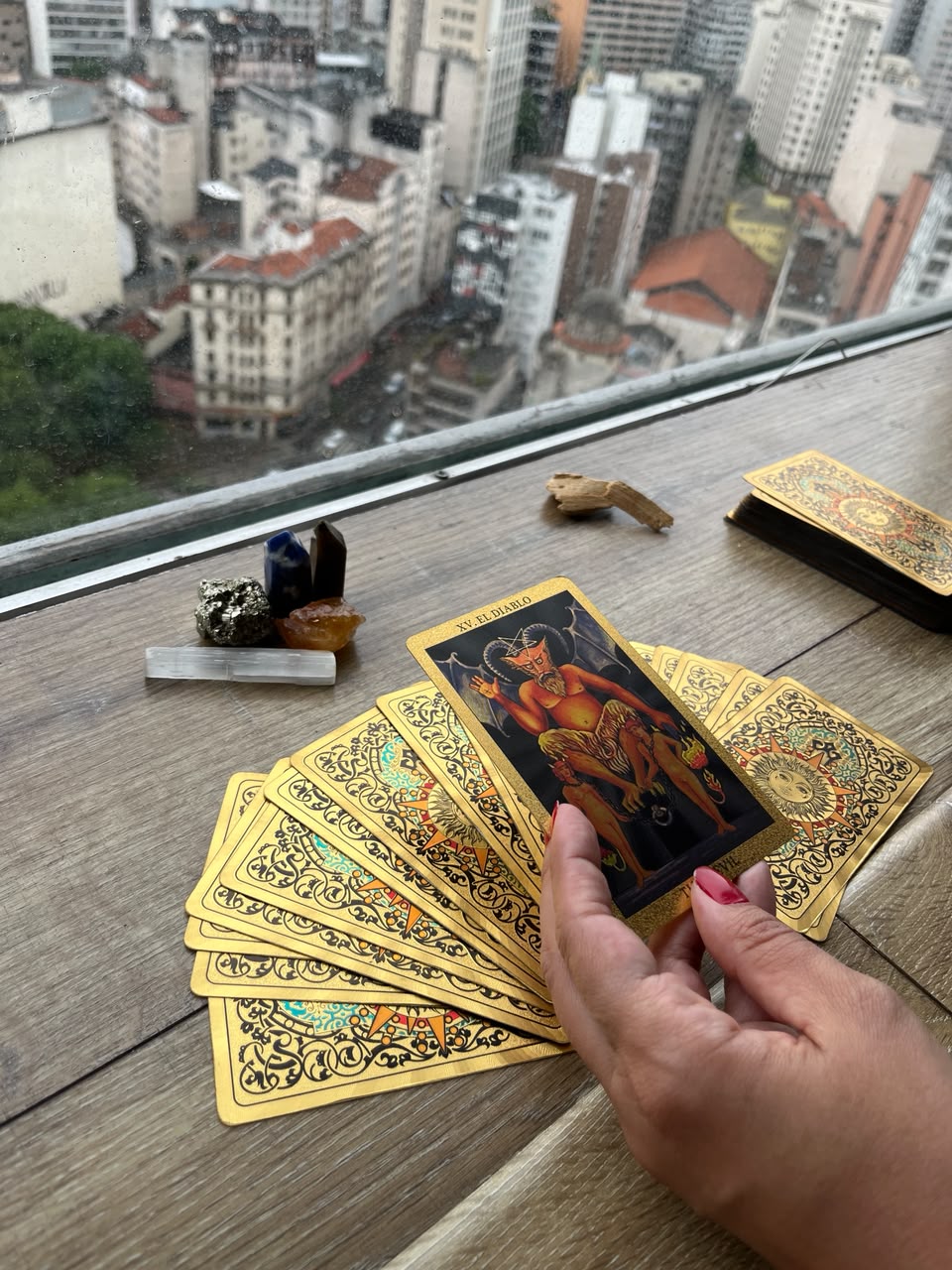 Cartas de tarot douradas seguradas em uma mesa com cristais e a cidade ao fundo